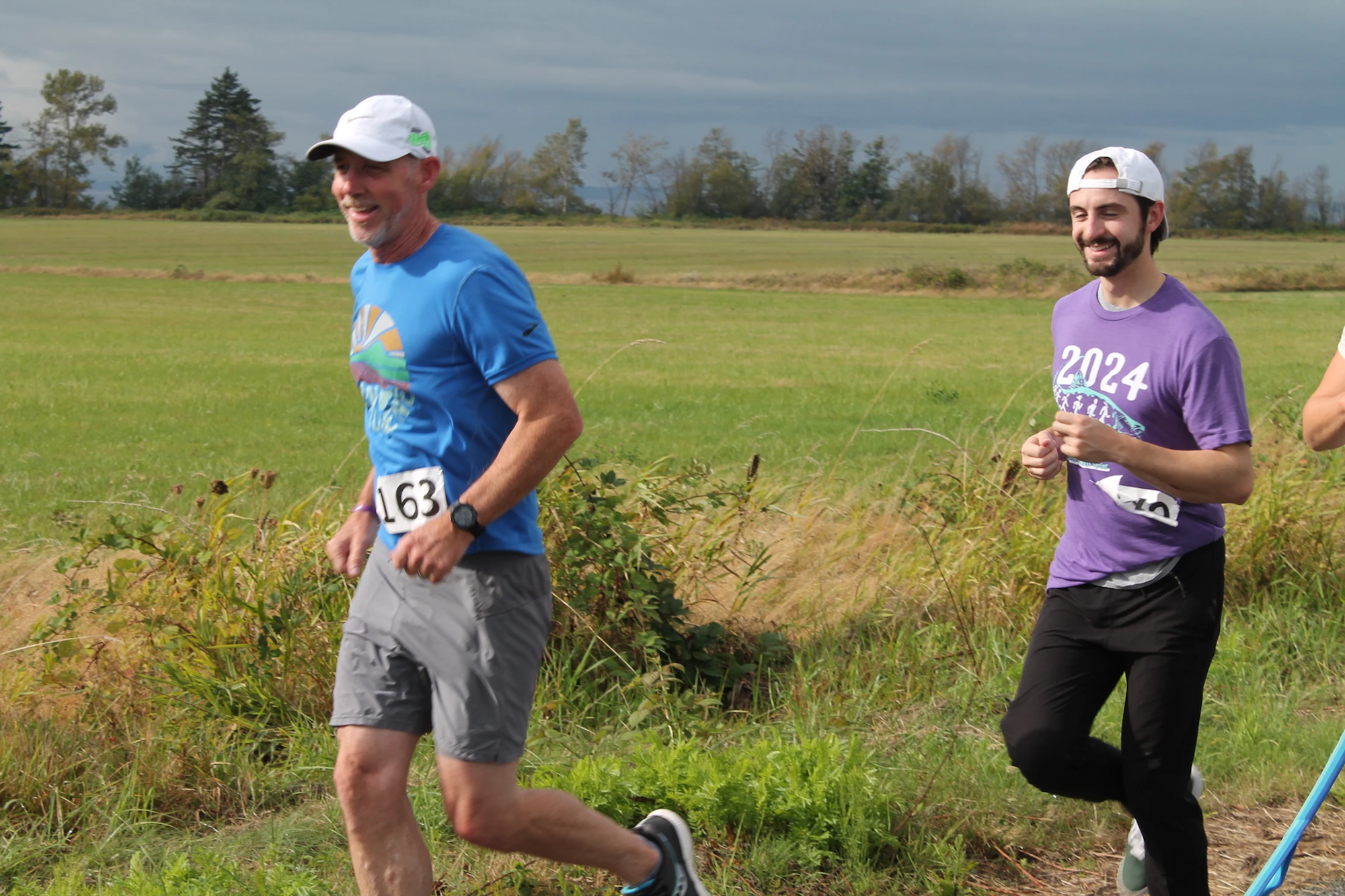 Two people are running outdoors with race numbers on their shirts, smiling in a green field.