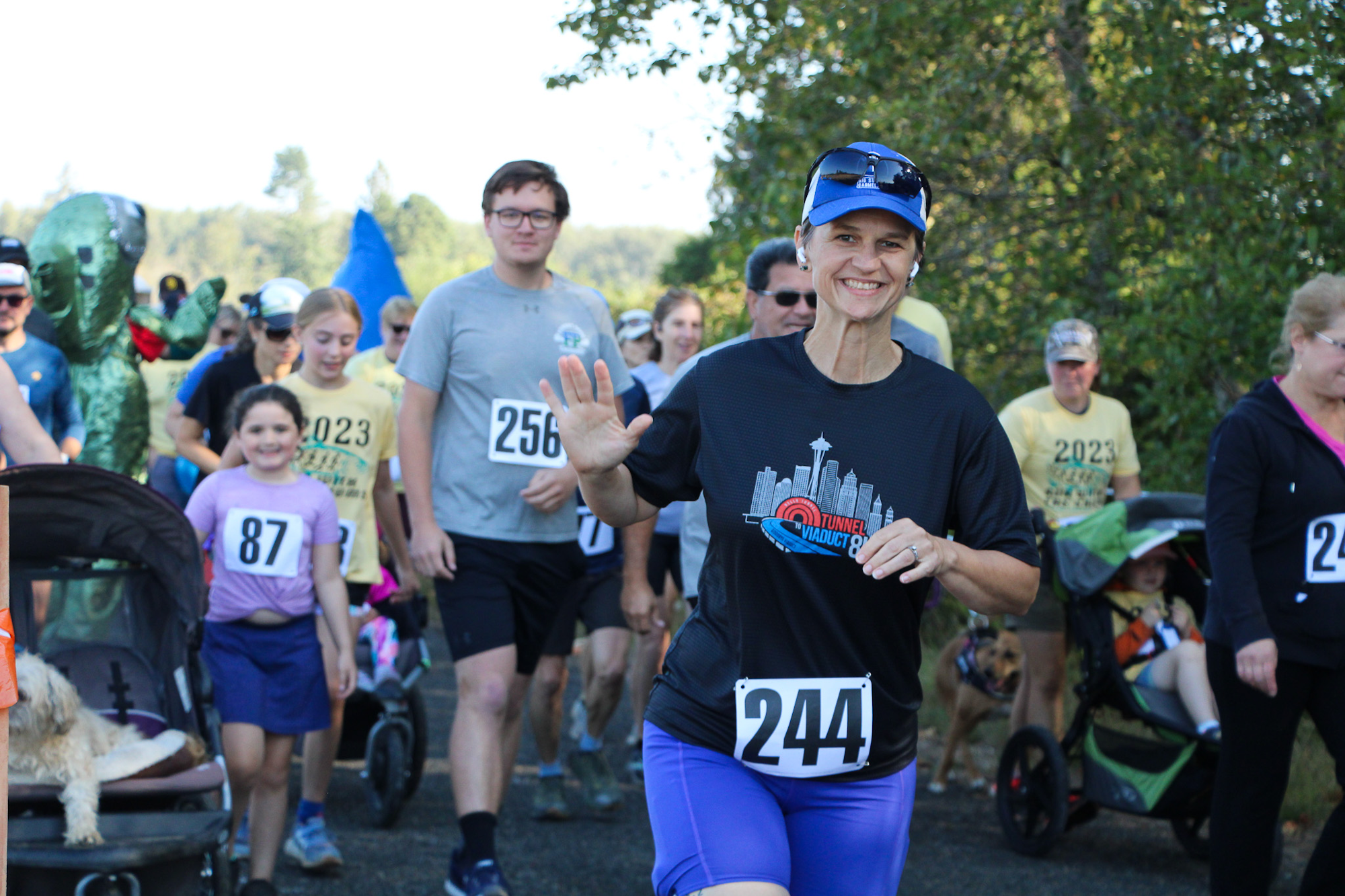 A group of people running in a race, wearing numbered bibs, with strollers and walkers in a cheerful outdoor setting.