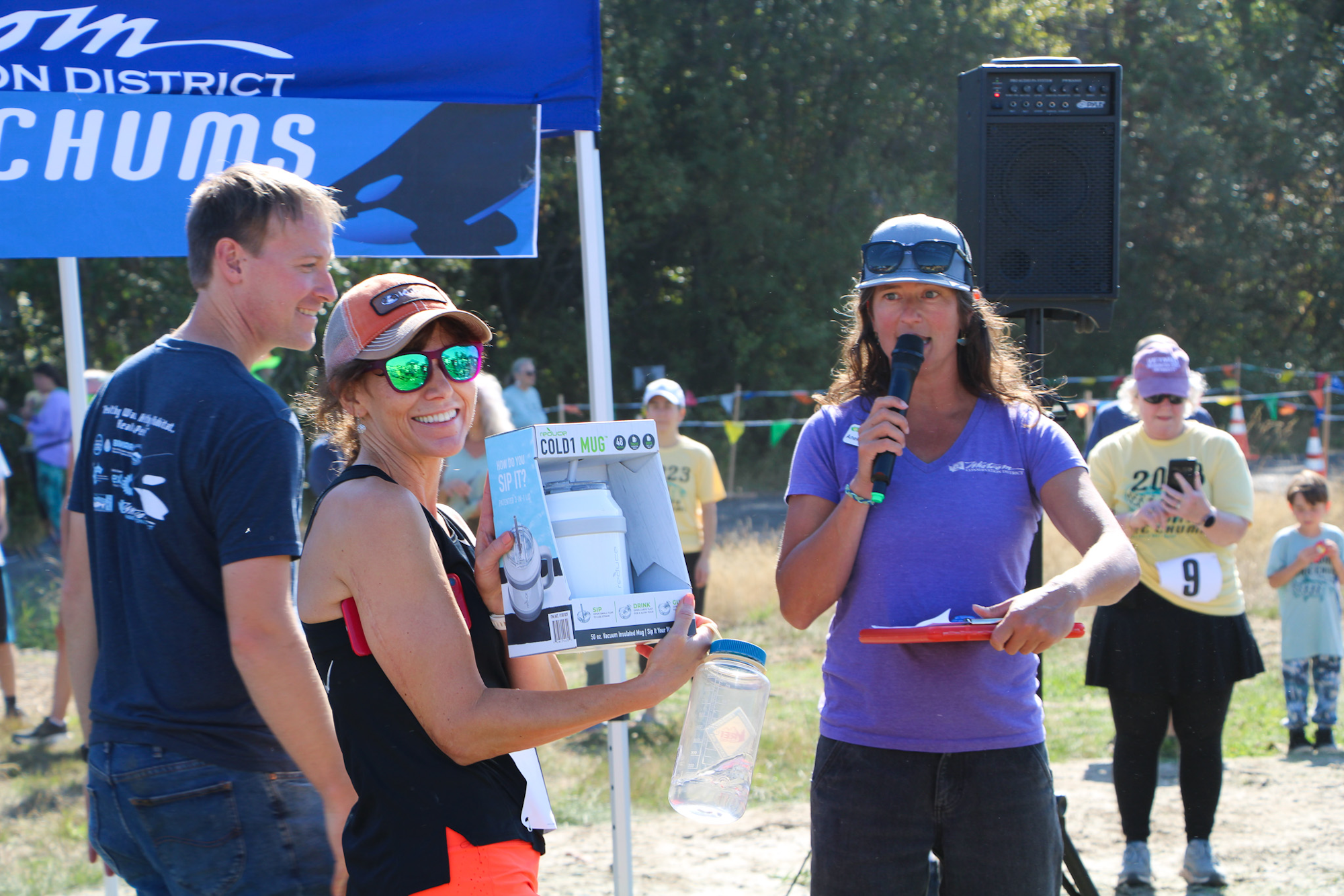 A woman holds a prize while another speaks into a microphone. People stand nearby at an outdoor event.