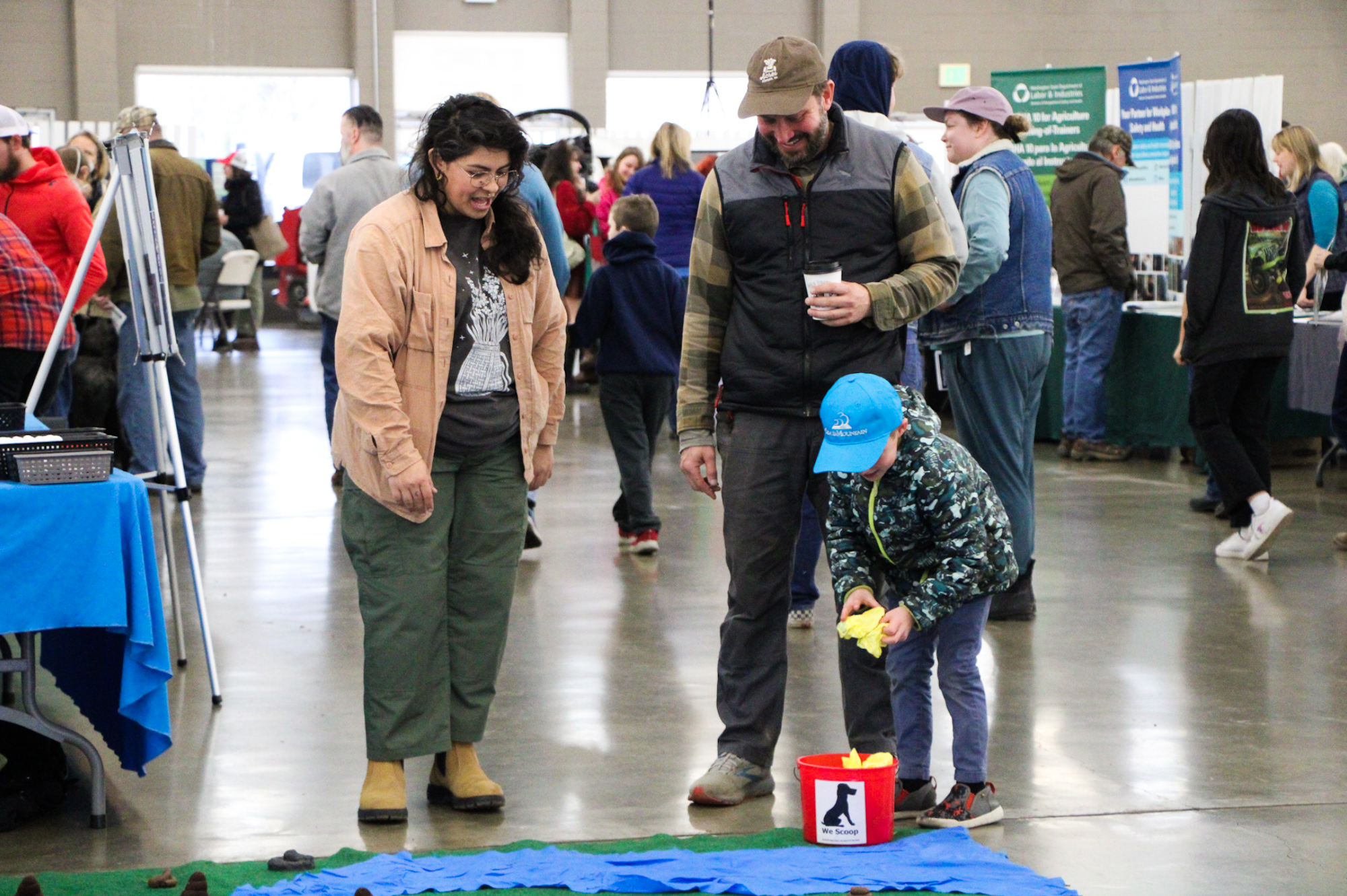 People at an indoor event watch a child play a game involving scooping, with a "We Scoop" bucket in the foreground.