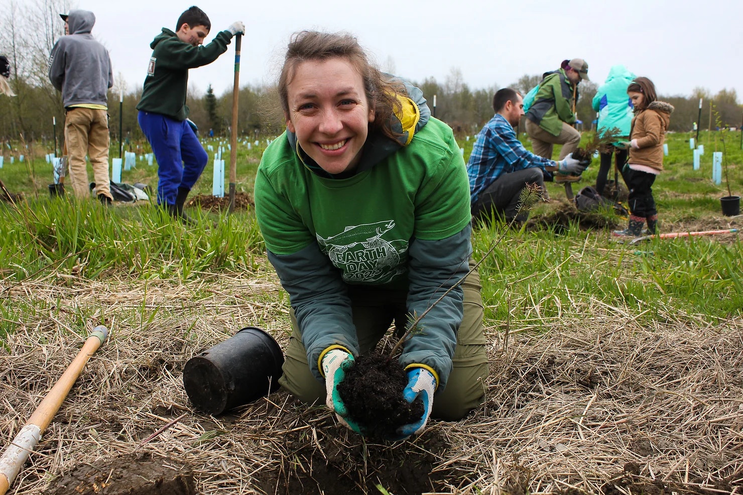 People planting trees outdoors, smiling woman in foreground holding a small plant.