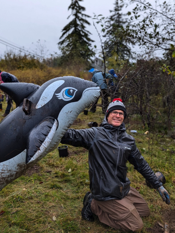 Person in outdoor gear smiling and holding an inflatable orca, with people and trees in the background.