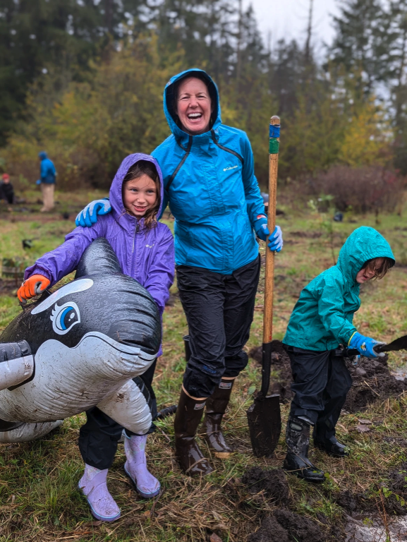 Three people in raincoats gardening; two kids and one adult smiling, with an inflatable orca.