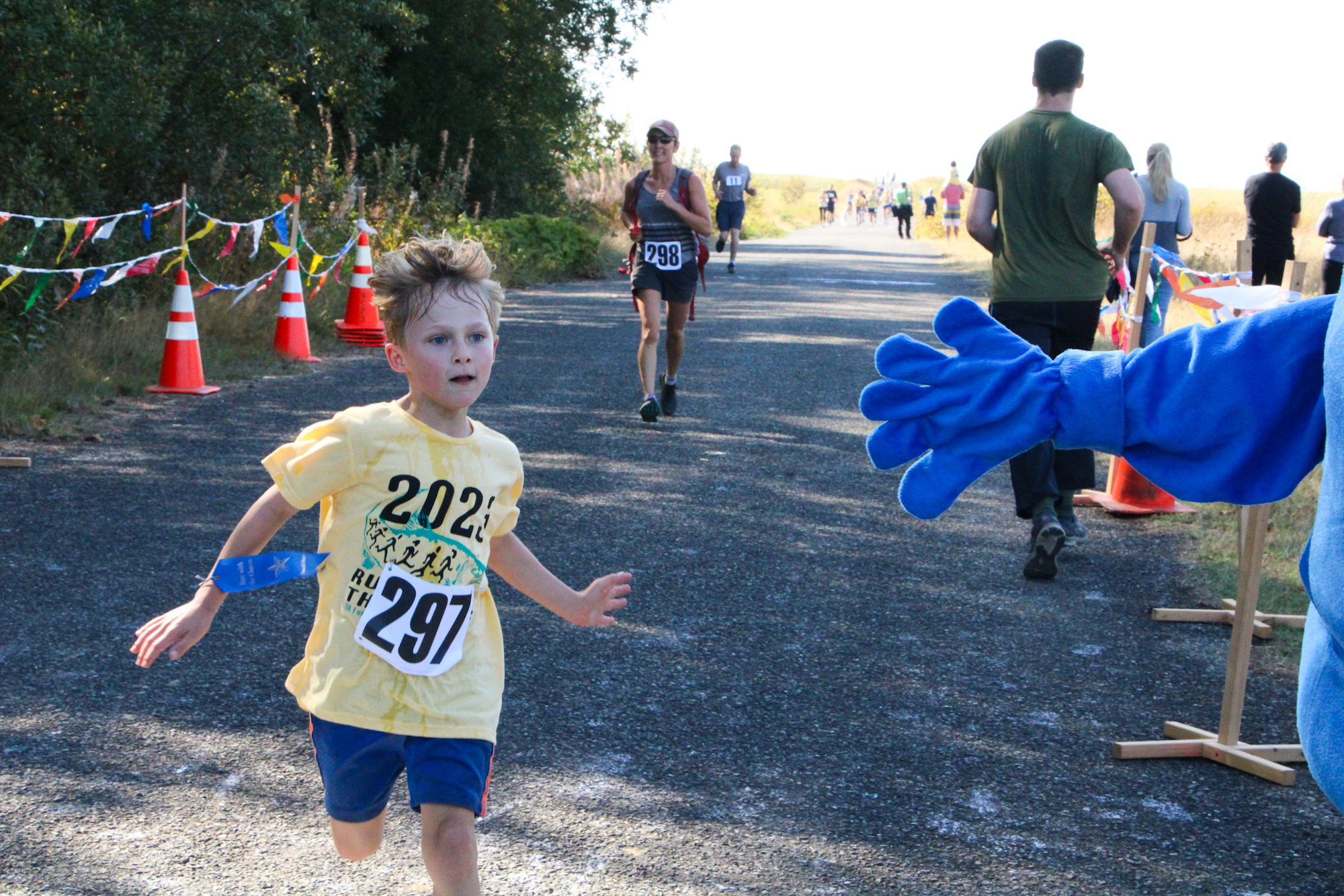 A young runner in a race, wearing a yellow shirt and numbered bib "297," approaches a person in a blue costume with an outstretched hand.