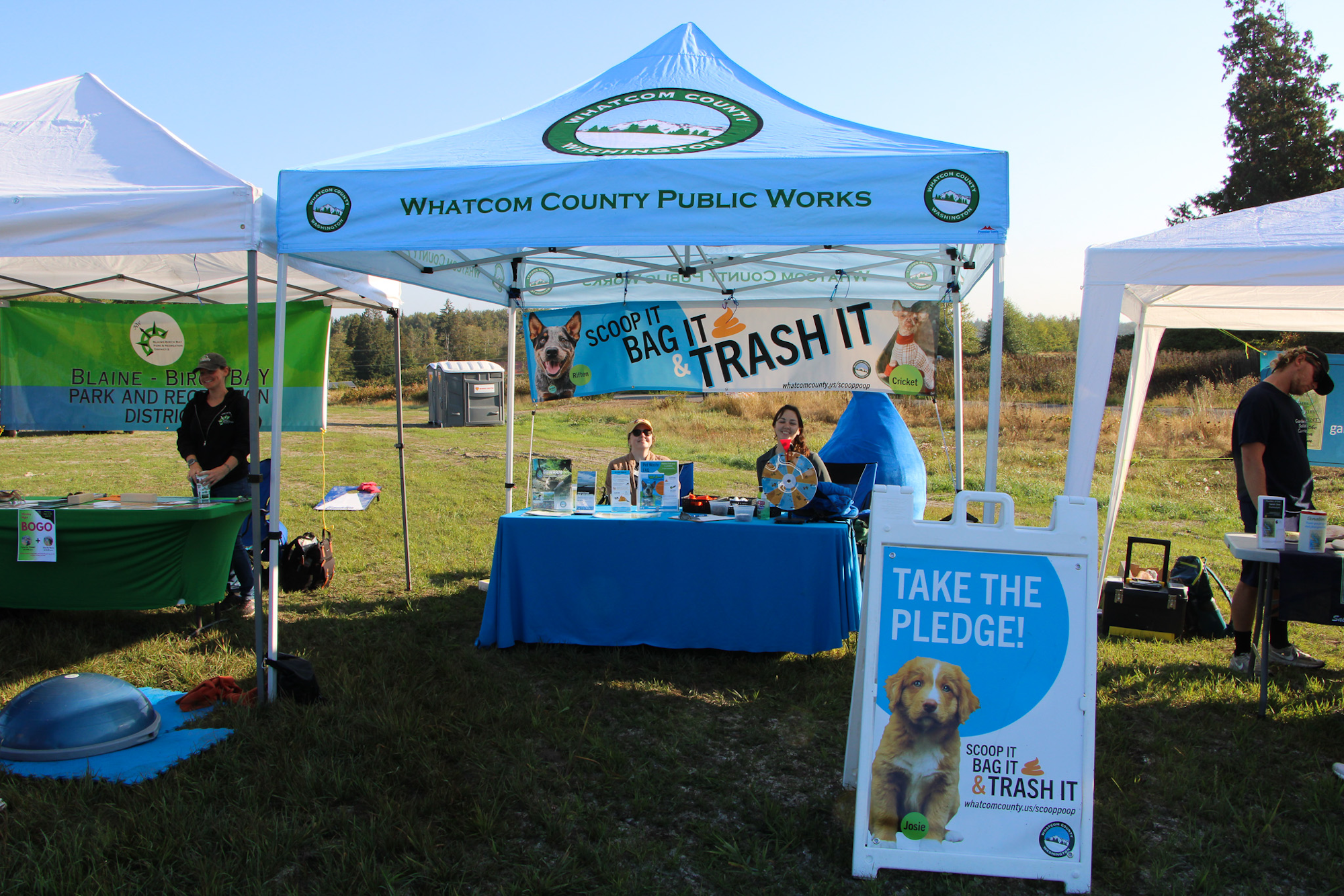 Outdoor event booths promoting pet waste disposal with a "Scoop it, Bag it, Trash it" message by Whatcom County Public Works.