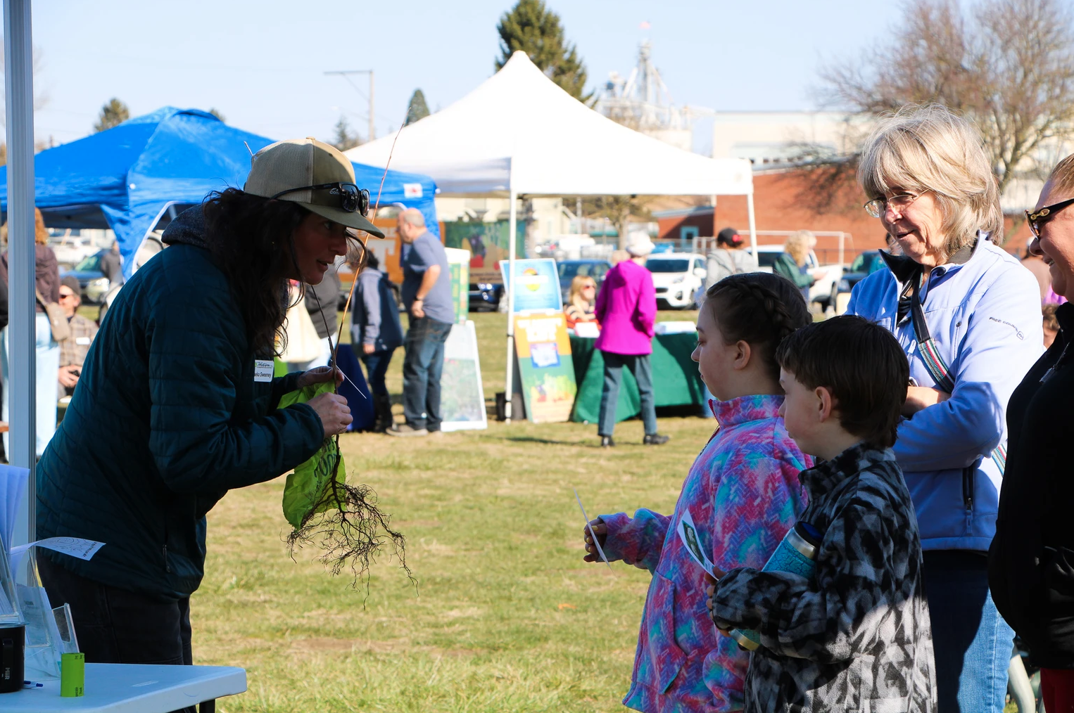 A woman shows plants to children at an outdoor event with booths and tents around.