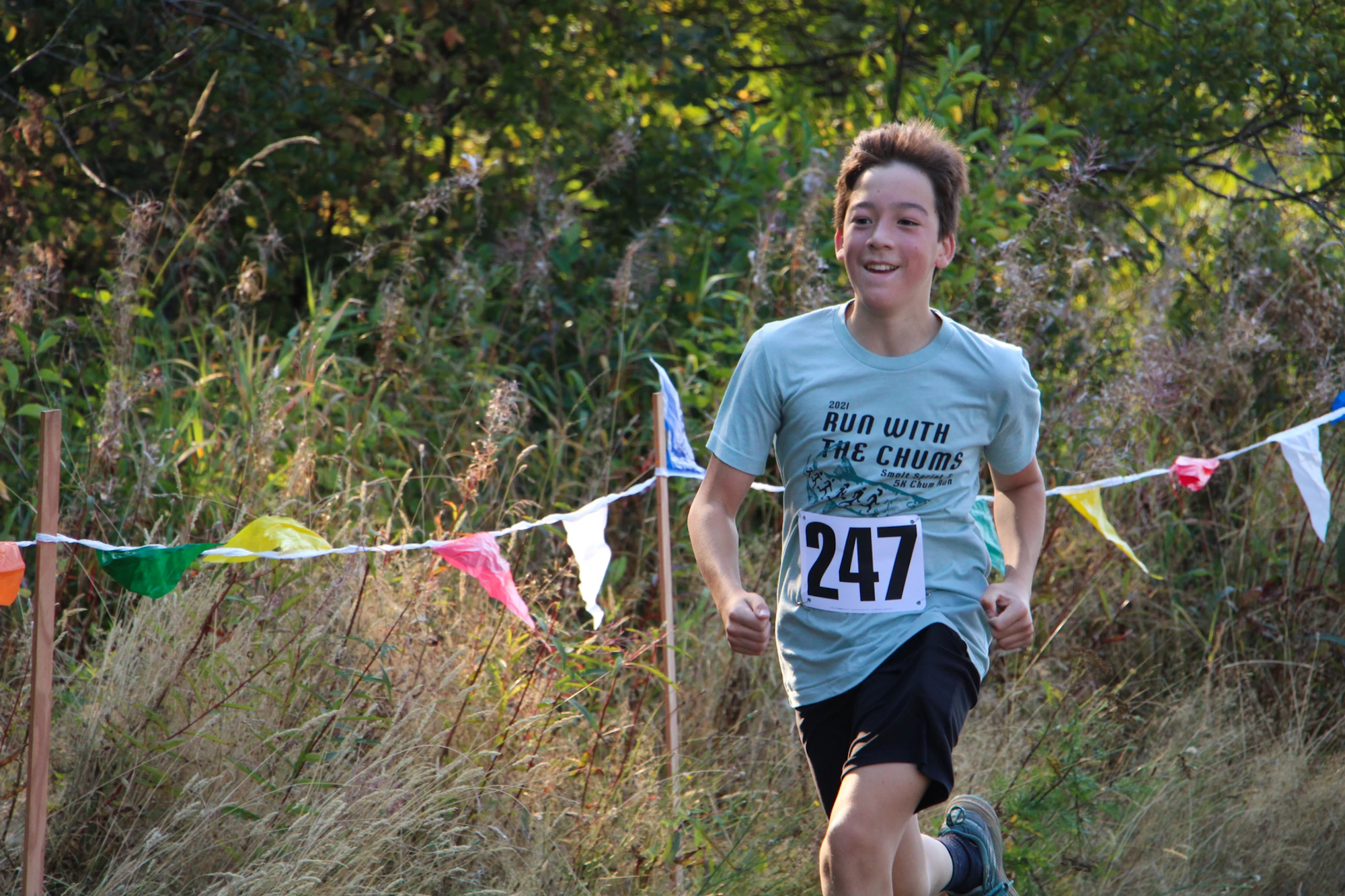 A person running a race through a grassy area, wearing a "Run with the Chums" shirt and race bib number 247.