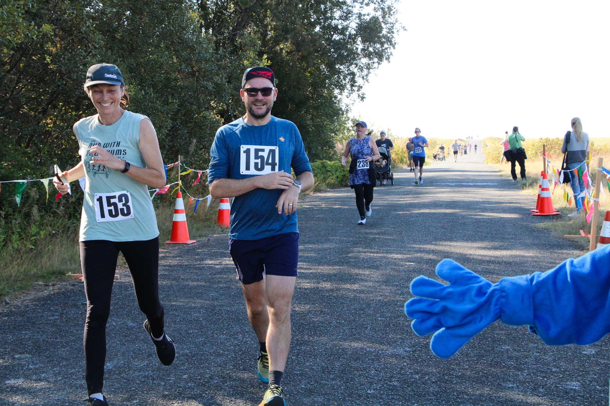 Runners with bibs 153 and 154 smiling during a race on a tree-lined path. Others follow behind, and a blue-costumed hand is visible.