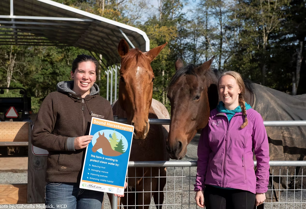 Two people with two horses, holding a "Sound Horsekeeping" sign, outdoors near a fenced area.