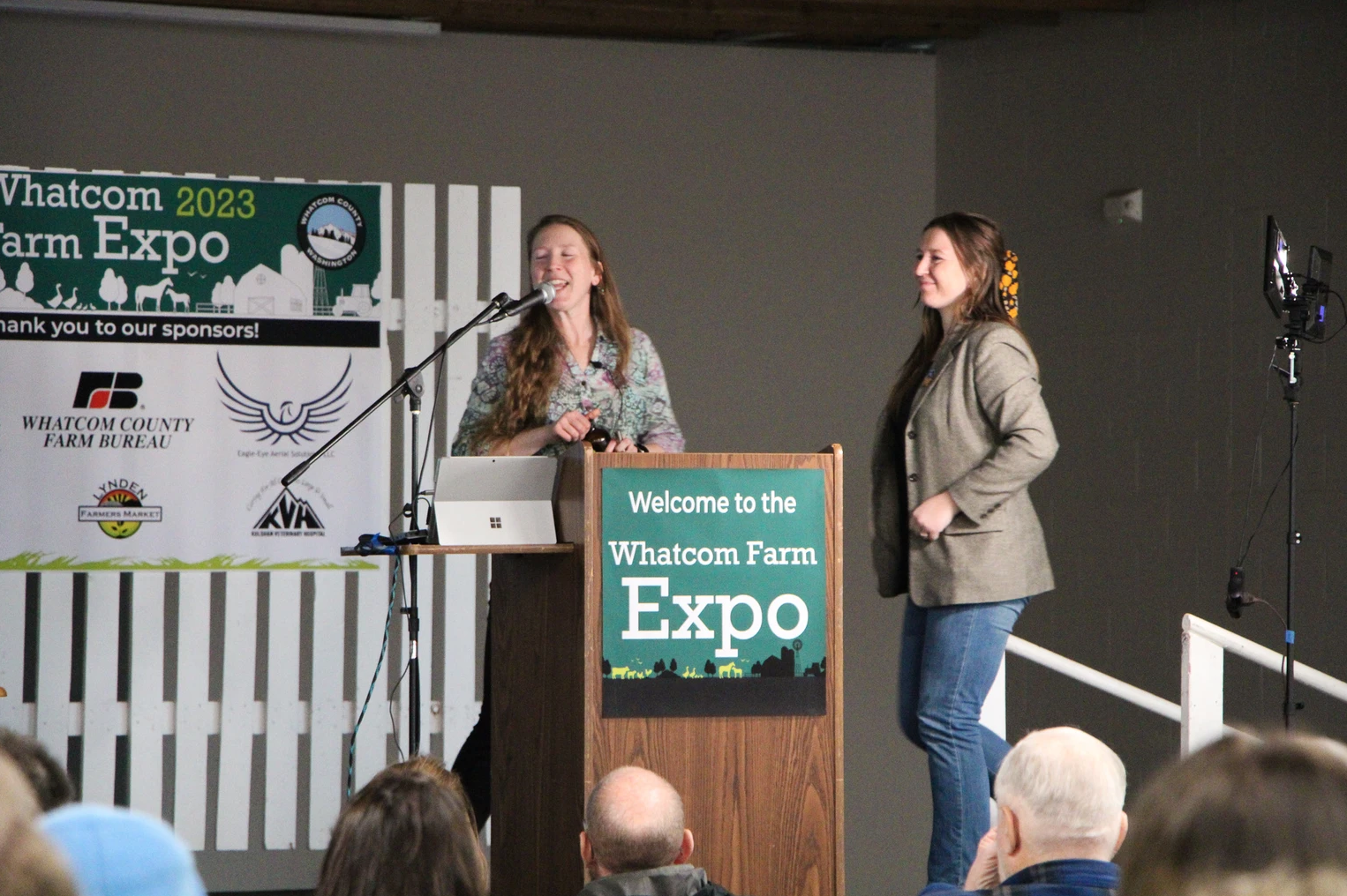 Two people speaking at the Whatcom Farm Expo, with a podium and banner in the background.