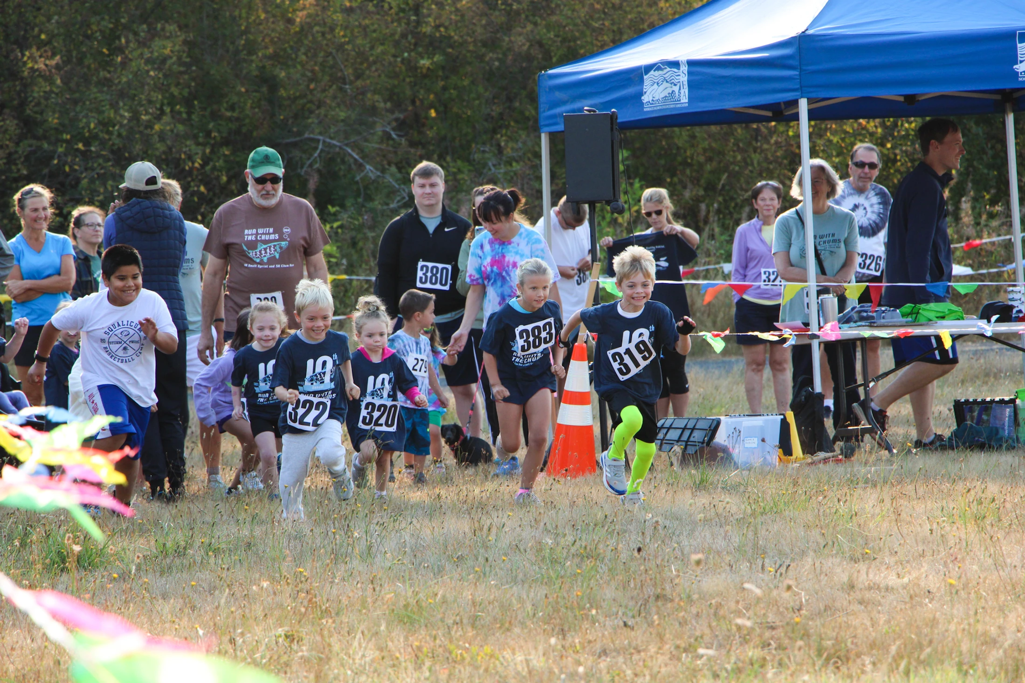 Children running a race with adults watching under a blue tent in a grassy area.