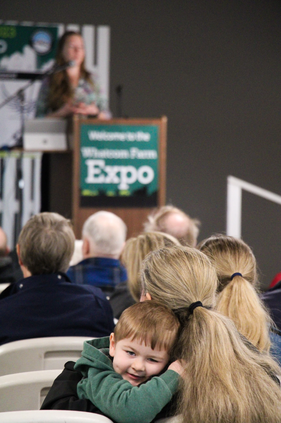 A child hugs an adult in an audience, with a speaker on stage at the Whatcom Farm Expo.