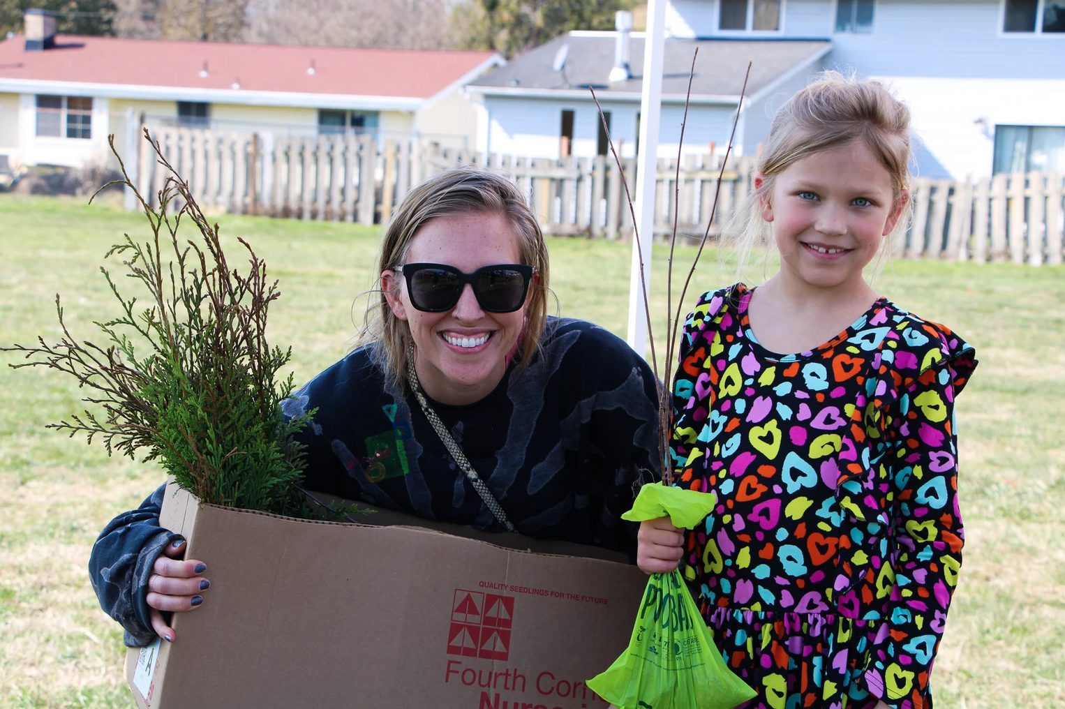 A woman and child smile outdoors; the woman holds a box with plants and the child holds a bag.
