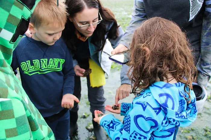 Children and adults examining objects, possibly shells, outdoors.