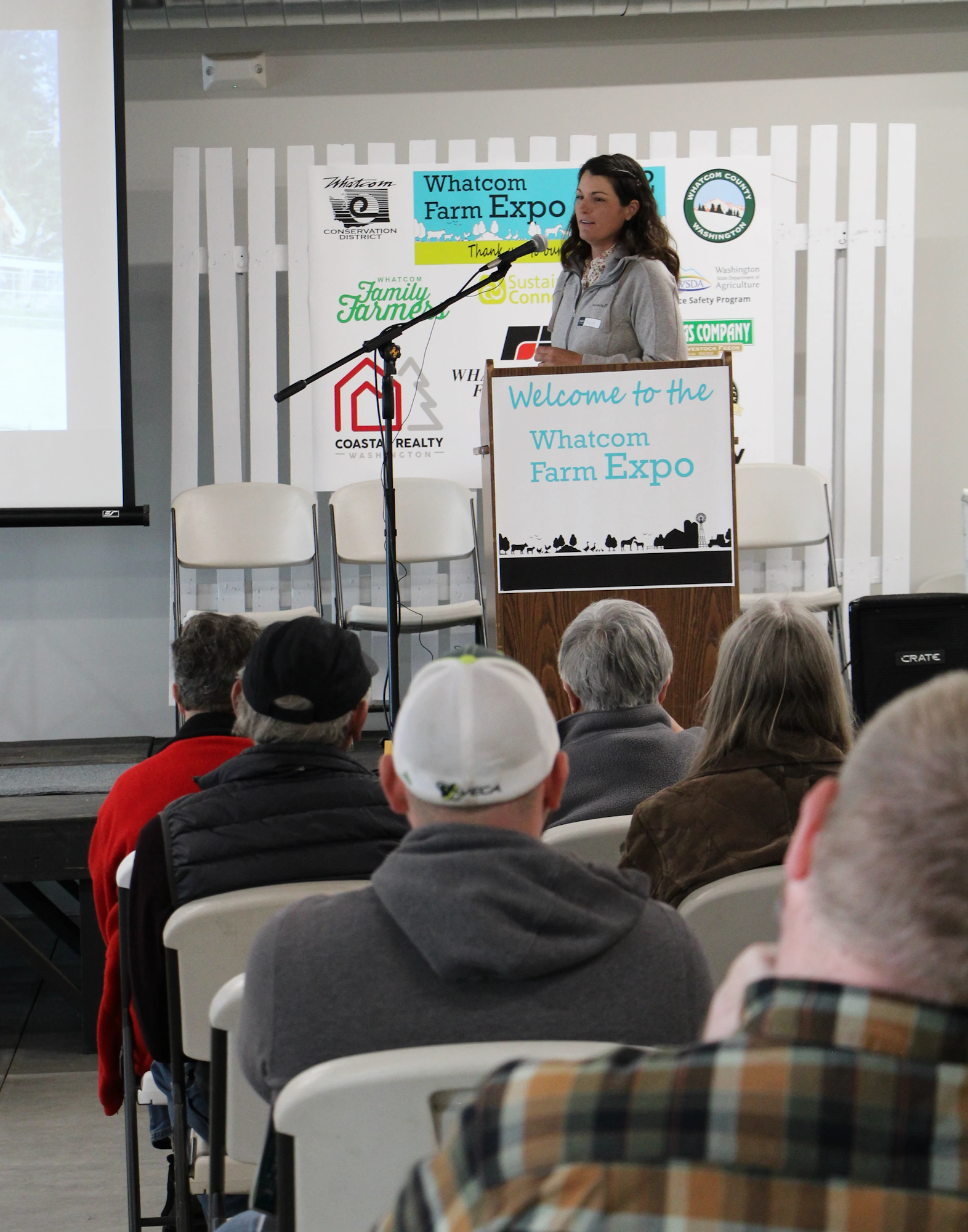 A woman speaking at a podium at the Whatcom Farm Expo, with an audience seated in front.