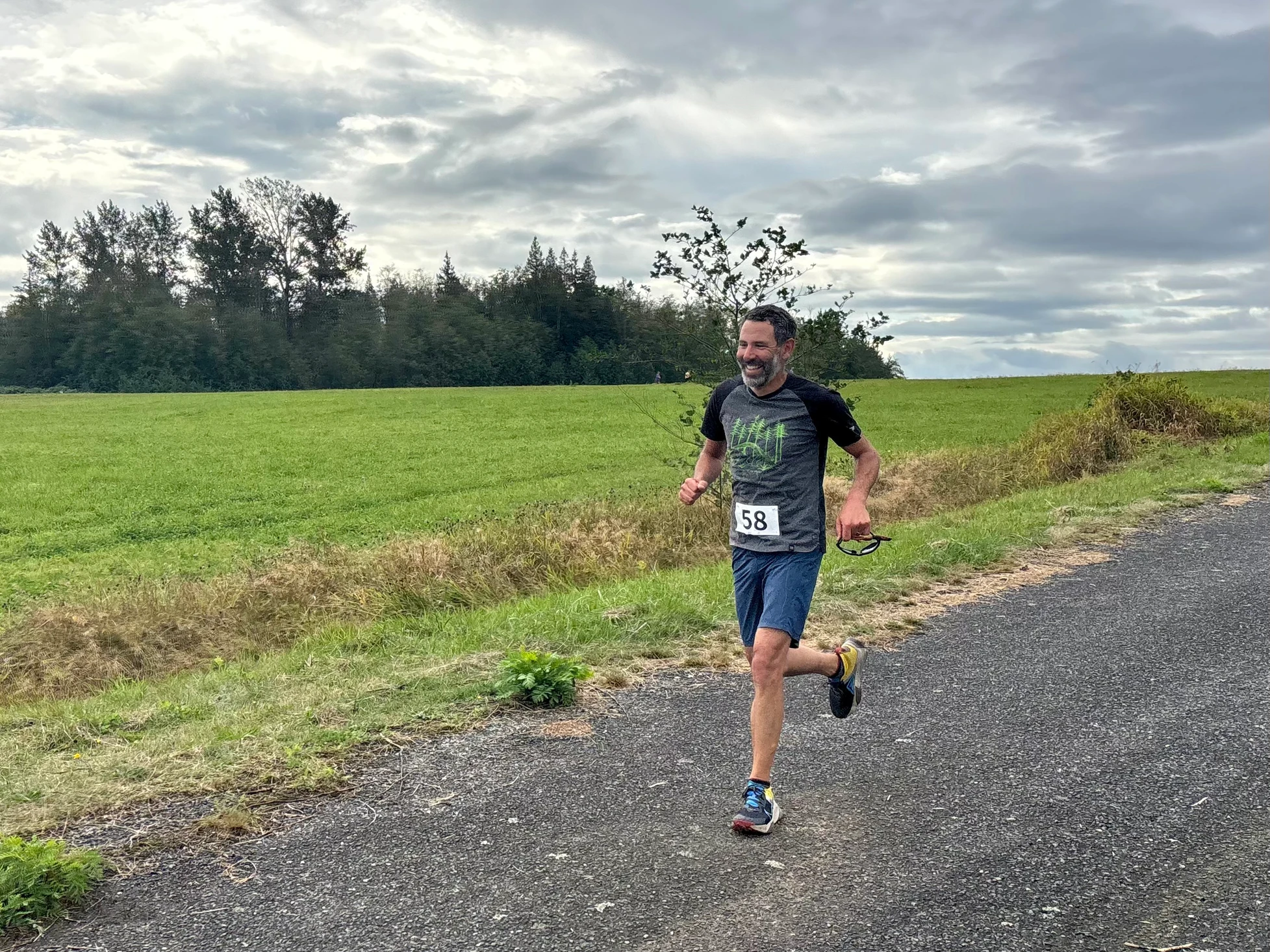 A person jogging on a rural path, wearing race number 58, holding sunglasses, with green fields and cloudy sky in the background.