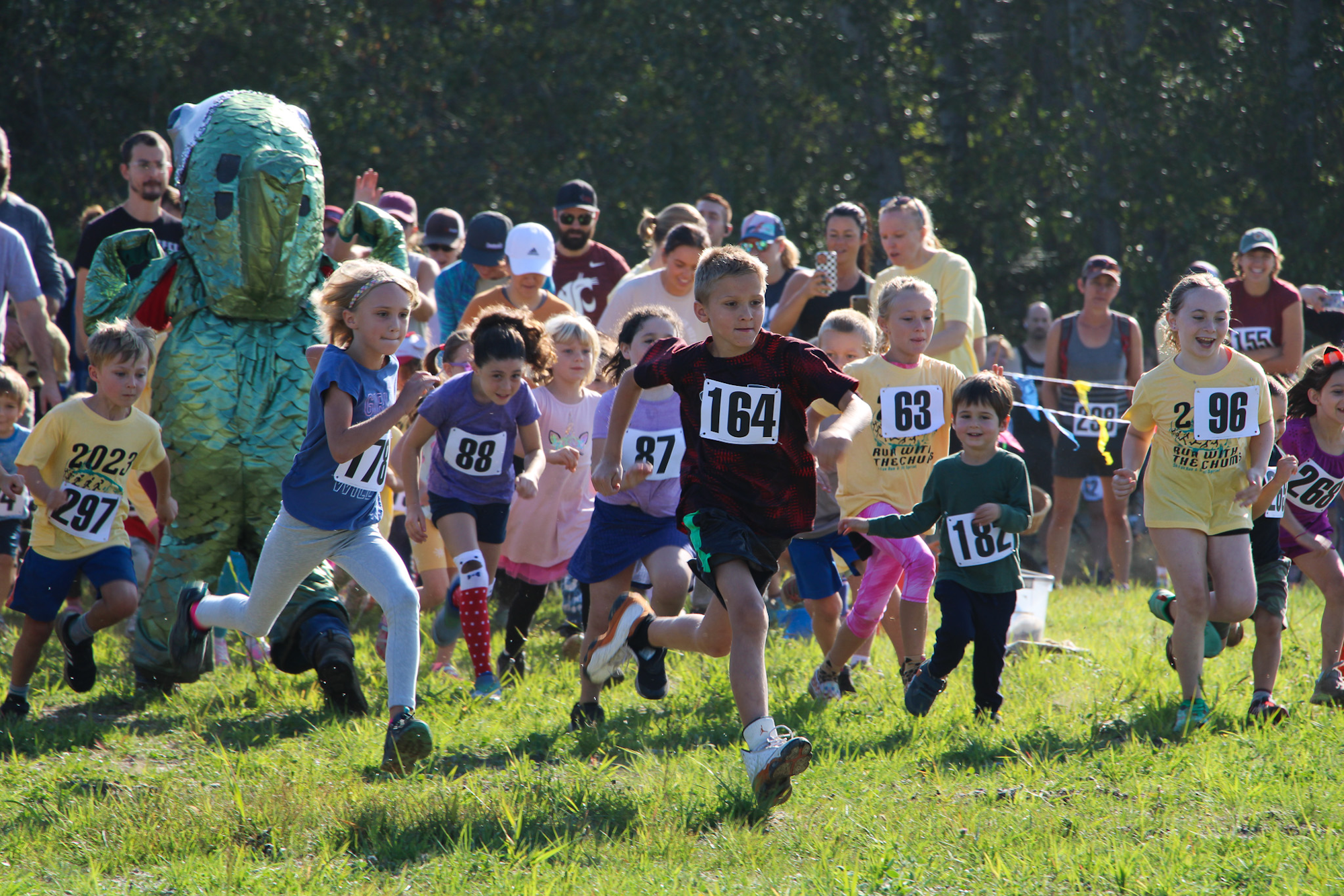 A group of children racing, wearing numbered tags, with a person in a dinosaur costume in the background.