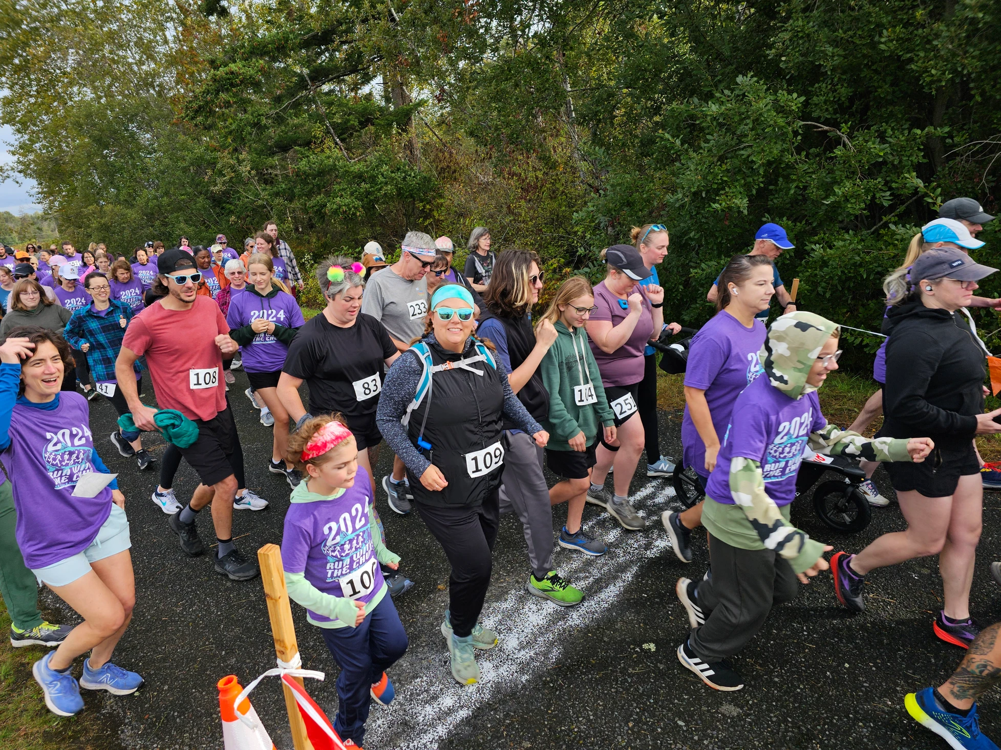 A group of people participating in a race or walk, wearing numbered bibs and various outfits, some with "2024" shirts.