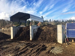A compost pile under a metal roof with a solar panel nearby in an outdoor setting.