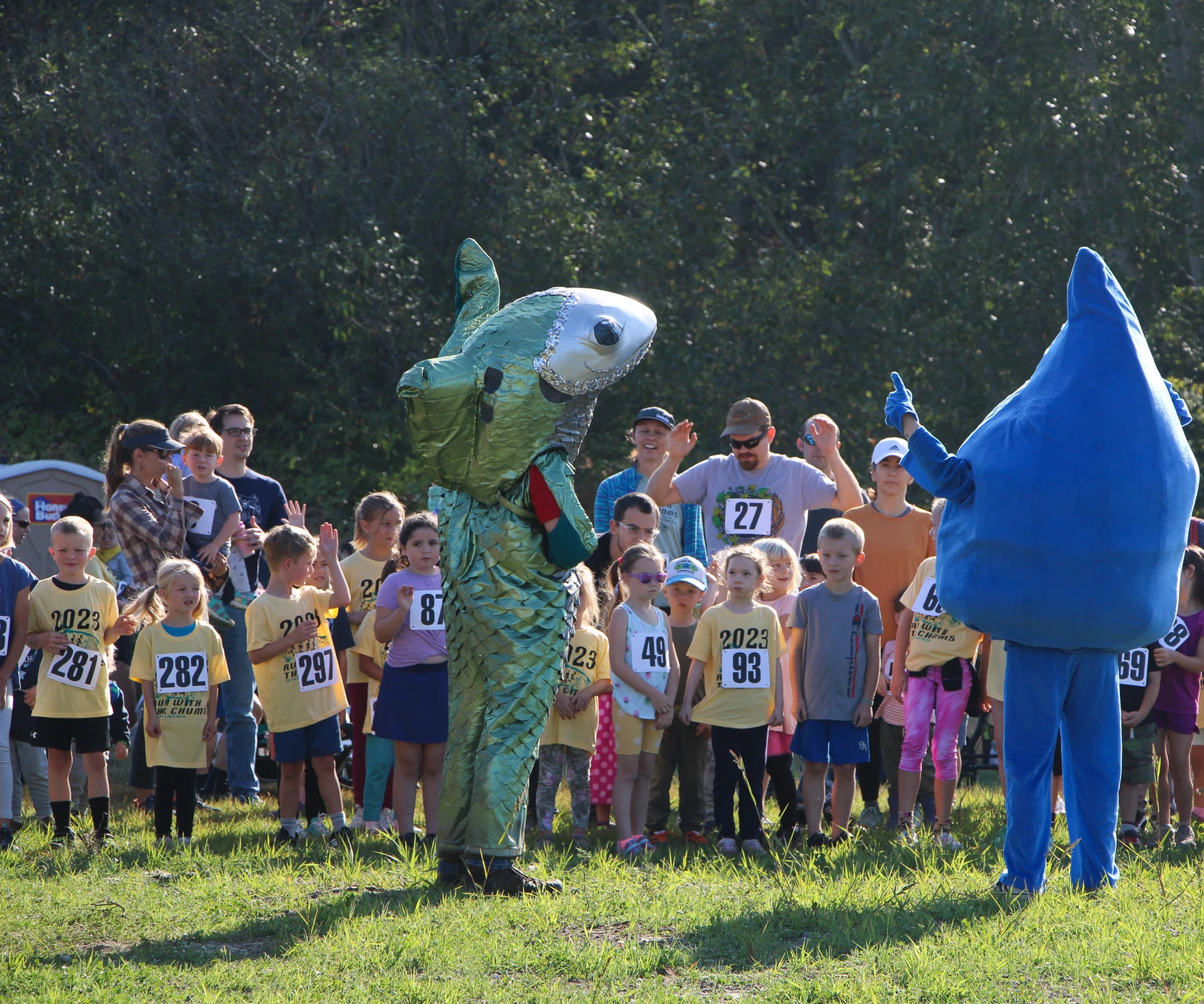 A group of children with bibs stand outdoors with two people in fish and raindrop costumes.