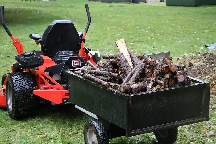 A lawn tractor with a trailer loaded with cut firewood on grass.