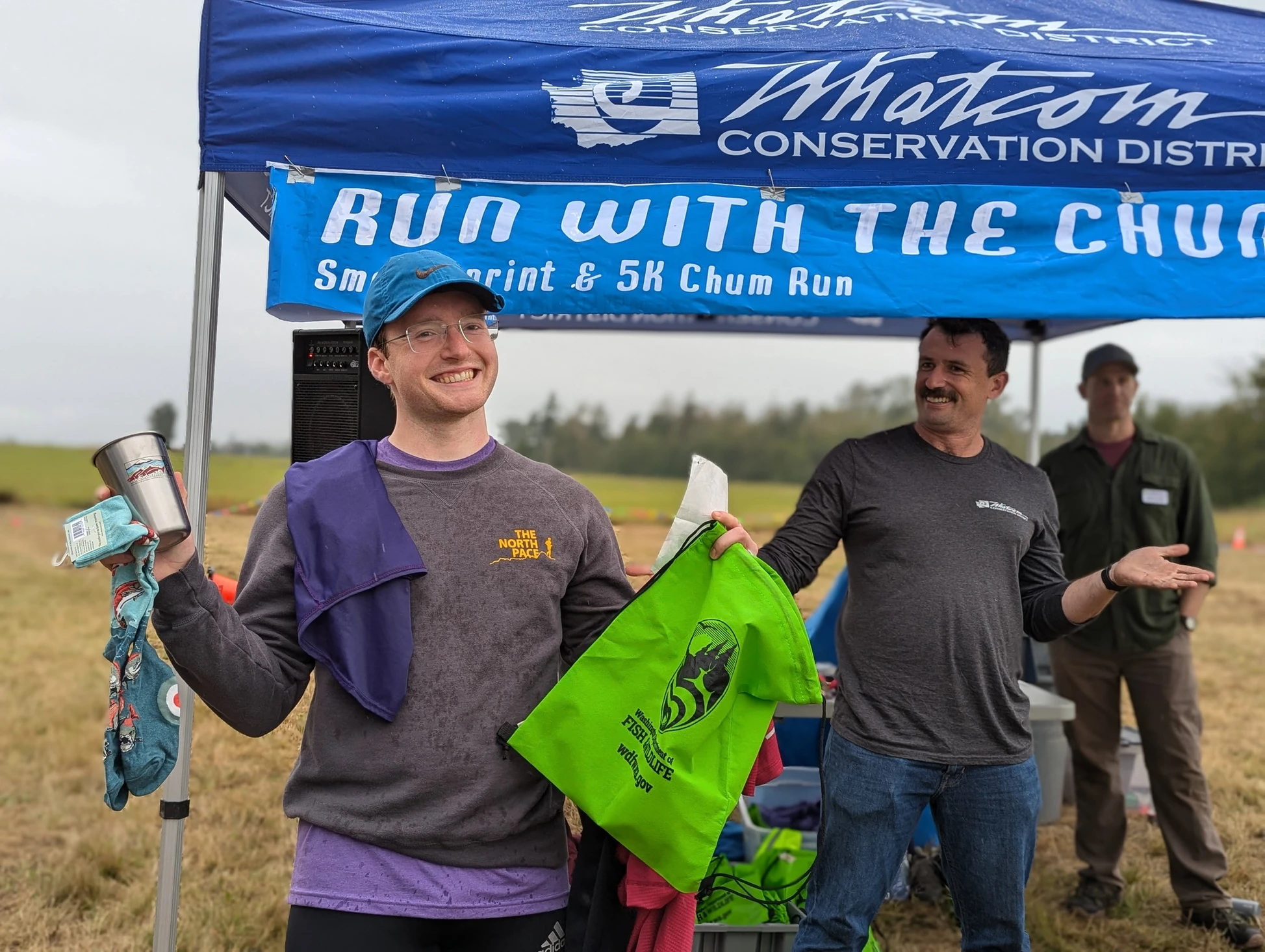 A person smiles holding prizes at a "Run with the Chums" event, under a blue tent with a conservation district banner.