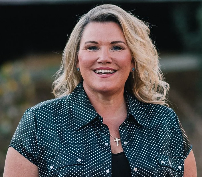 A person with blonde hair smiles, wearing a black polka dot shirt and a cross necklace.