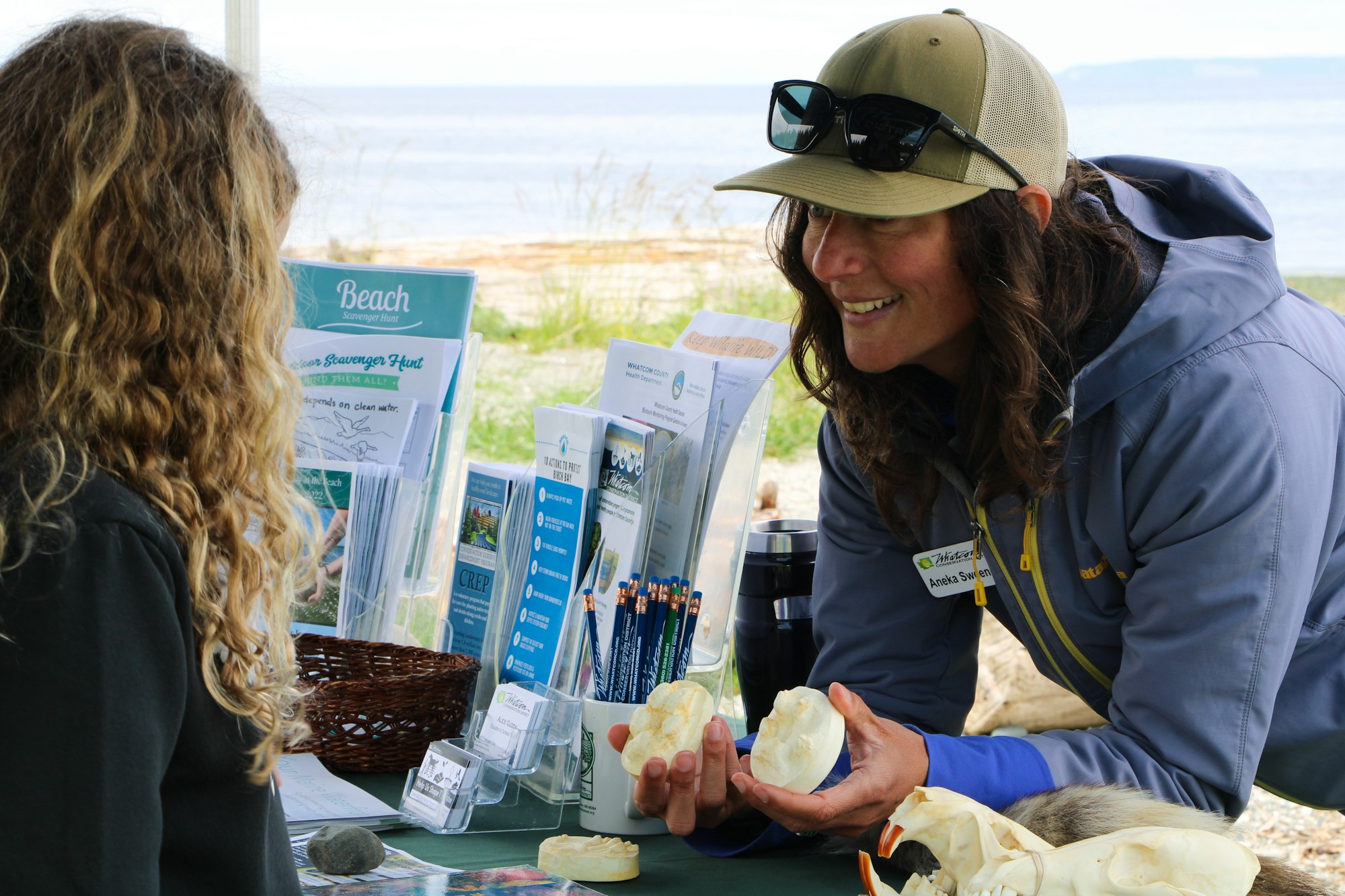 A woman shows animal skulls to a girl at an outdoor educational booth by the beach.