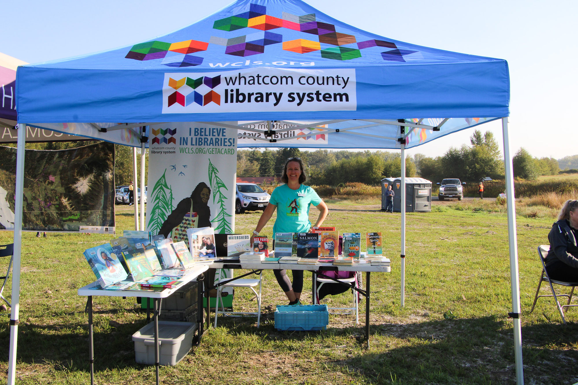 A library booth with books, a person, and a sign for Whatcom County Library System under a blue canopy in an outdoor setting.
