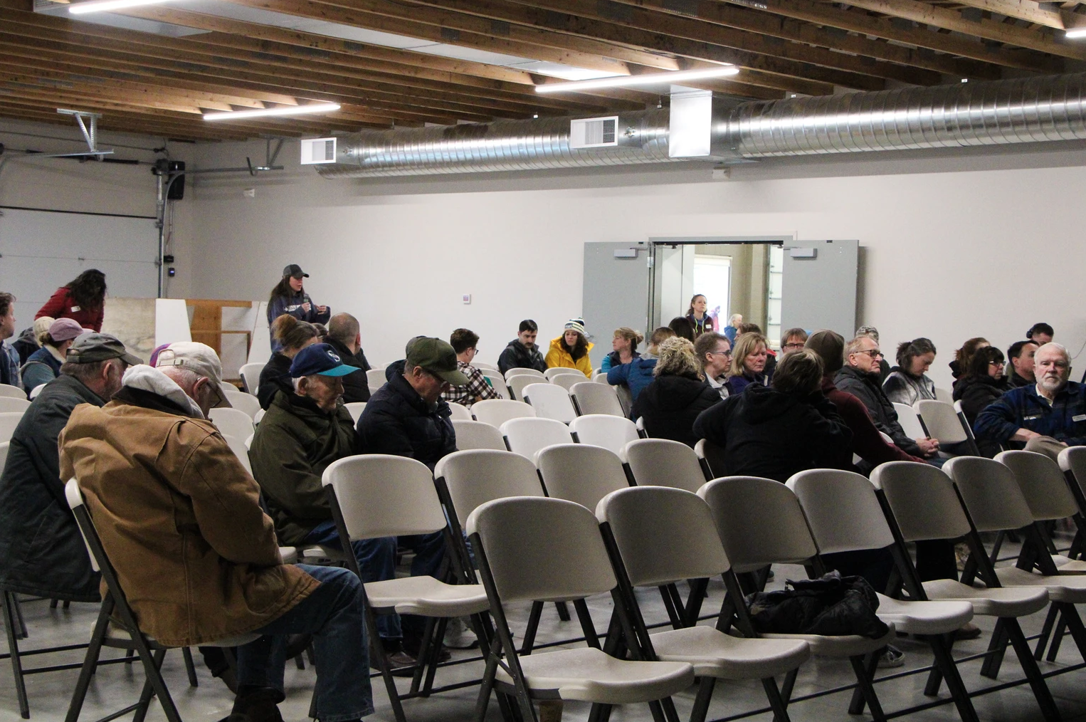 A small group of people seated in a room with rows of chairs, some talking, in an indoor setting.