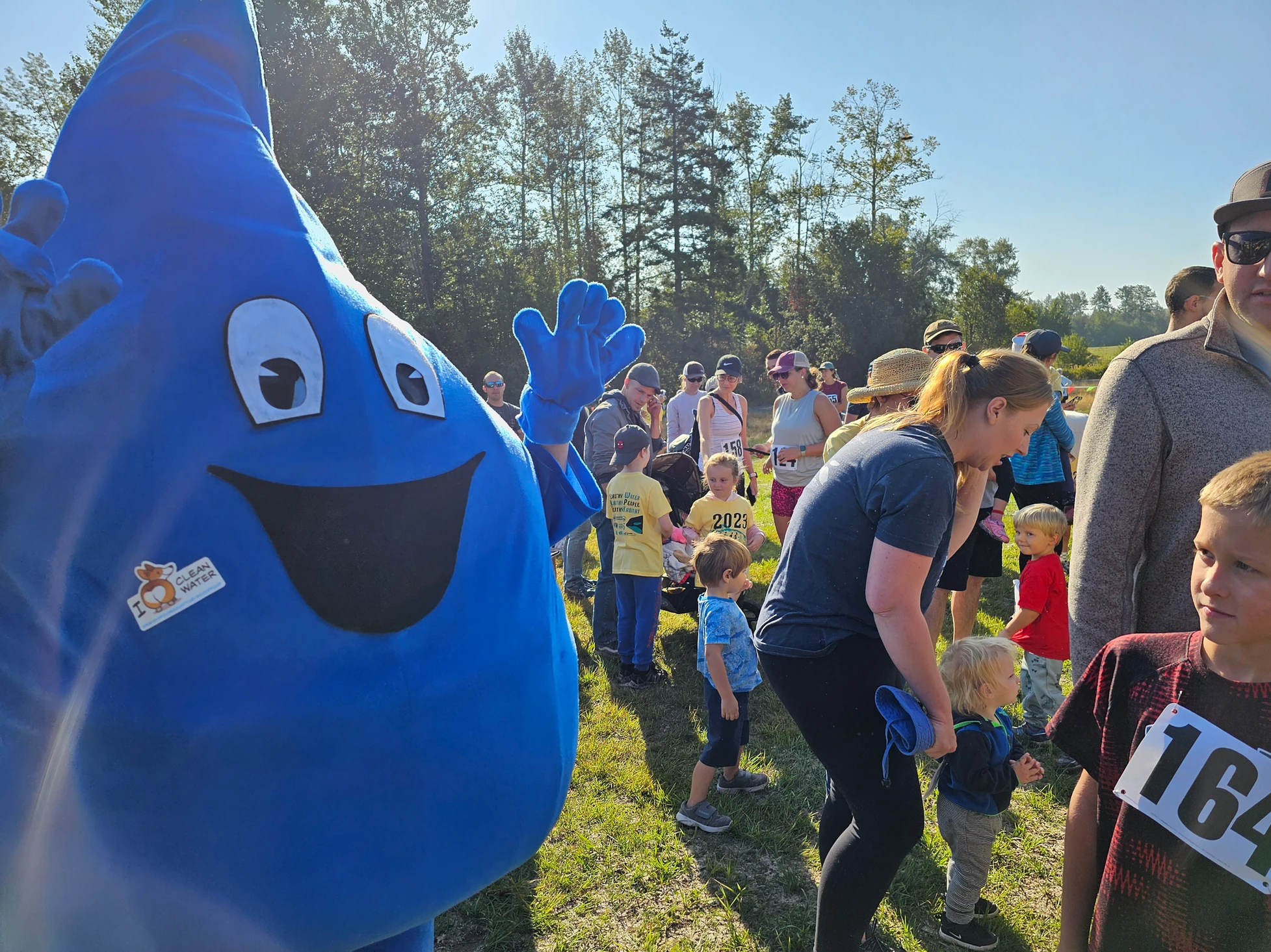 A blue mascot with a "I ❤️ Clean Water" sticker stands among a crowd, including kids with race numbers in a sunny outdoor setting.