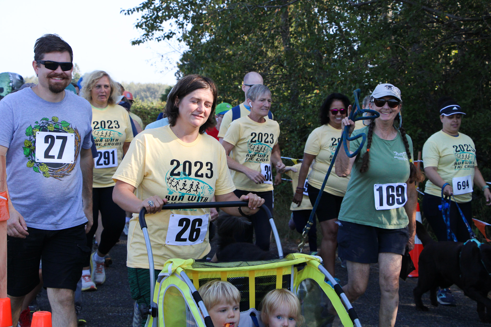 Group participating in a 2023 run event, wearing numbered bibs and t-shirts, with a stroller and a dog among them.