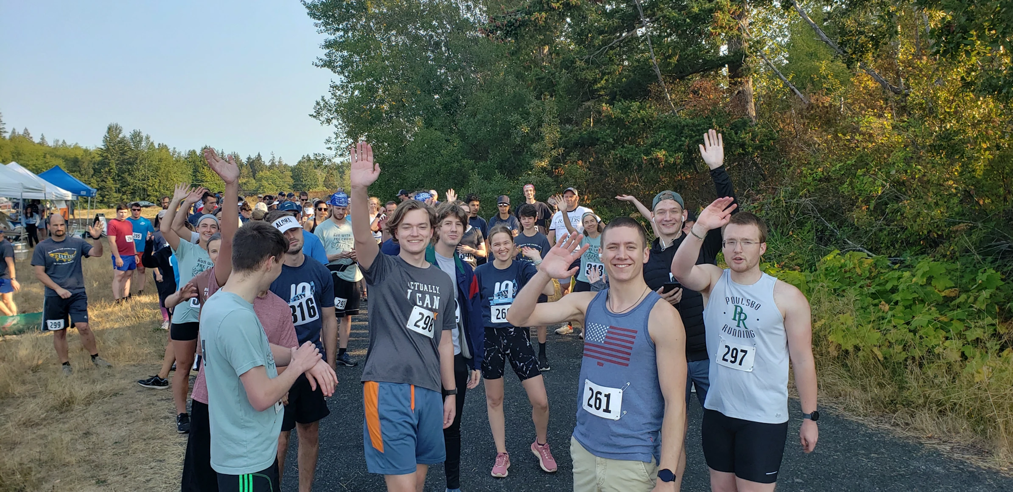 Group of people with race numbers, waving, outdoors with trees in the background.