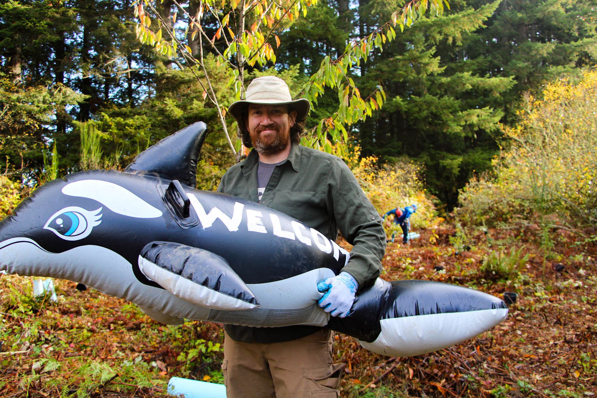A person outdoors holding an inflatable killer whale with "Welcome" on it, set against a forest backdrop.