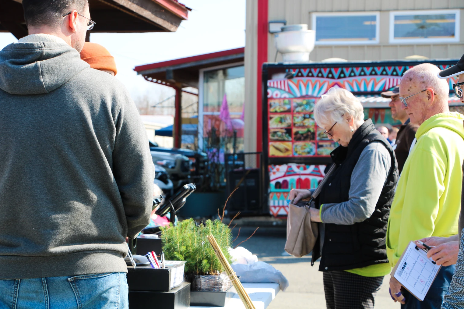 People stand in line at an outdoor market, one woman holding a bag. In the background, a colorful food truck.
