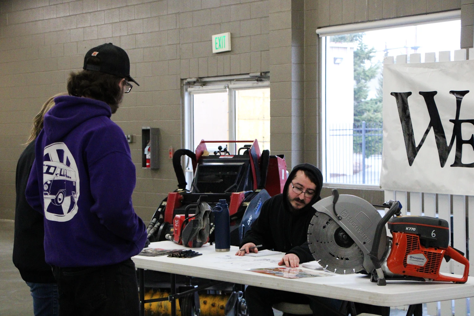 Two people at a table with a saw, safety gear, and materials, indoors.