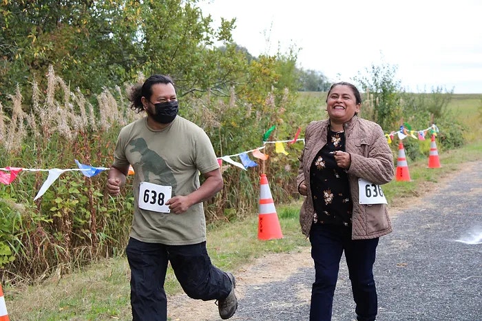 Two people running a race outdoors, both smiling, with number bibs, near traffic cones and colorful flags.