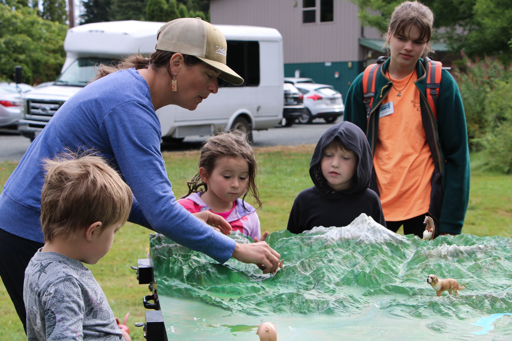 A group of children and an adult interact with a green landscape model outdoors, observing and learning.