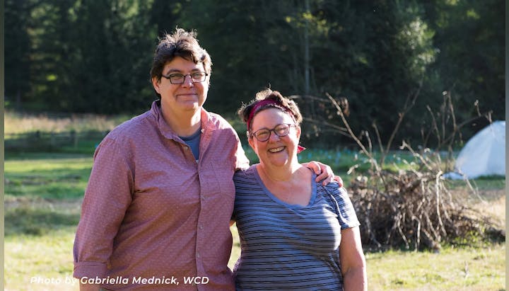 Two people smiling outdoors in a sunny field with trees in the background.