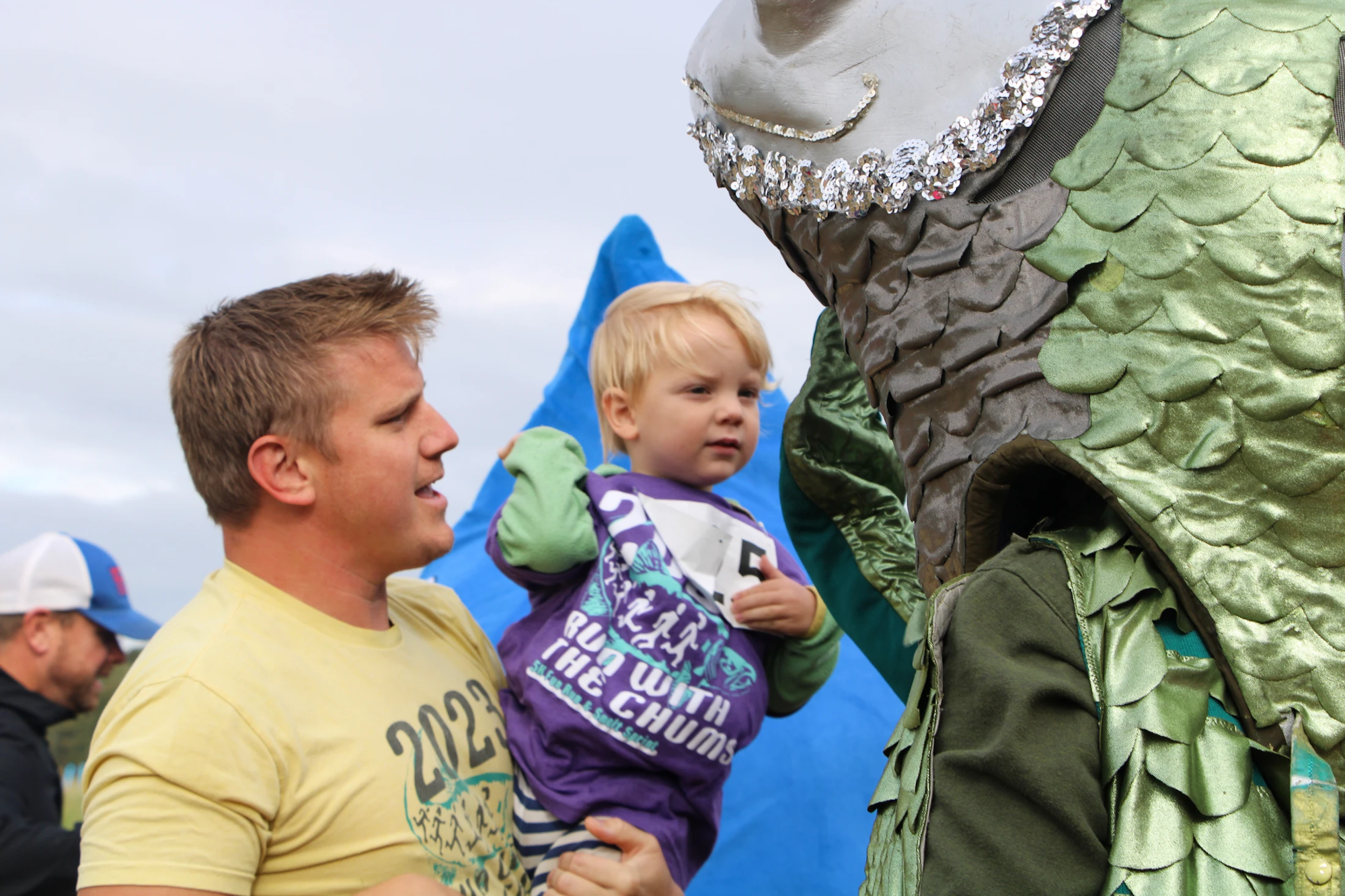 A man holds a child wearing a "Run with the Chums" shirt, standing next to a person in a dragon or fish costume.