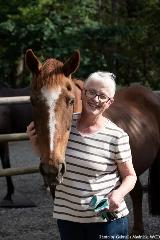 A person smiling next to a horse, holding its head gently, with a natural background.