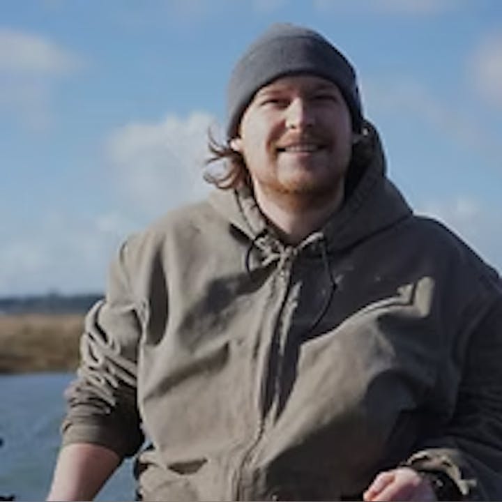 A person in a gray hoodie and beanie smiling outdoors near a body of water.