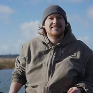 A person in a gray hoodie and beanie smiling outdoors near a body of water.