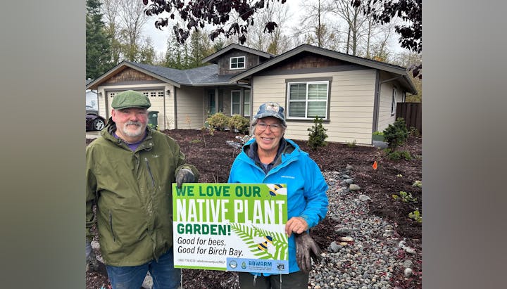 Two people in front of a house hold a sign promoting a native plant garden for bees and Birch Bay.
