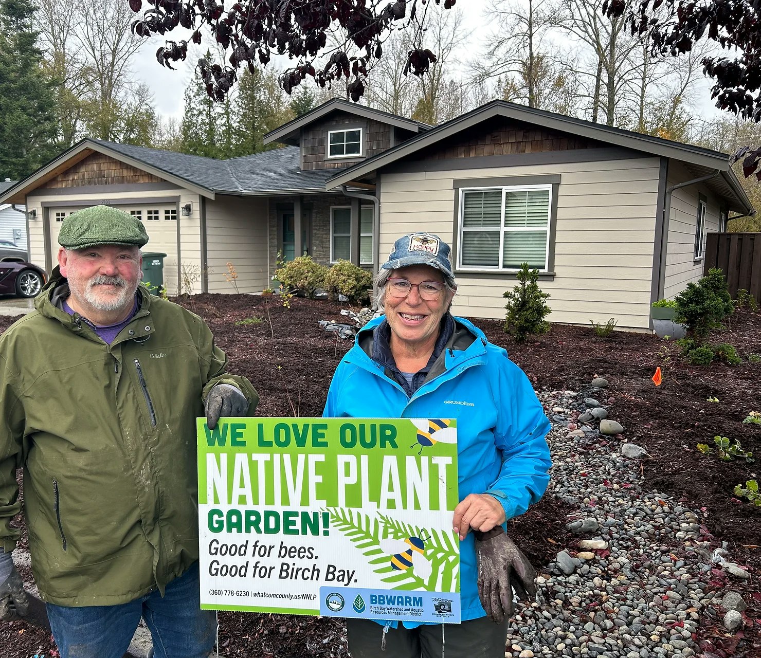Two people in front of a house hold a sign promoting a native plant garden for bees and Birch Bay.