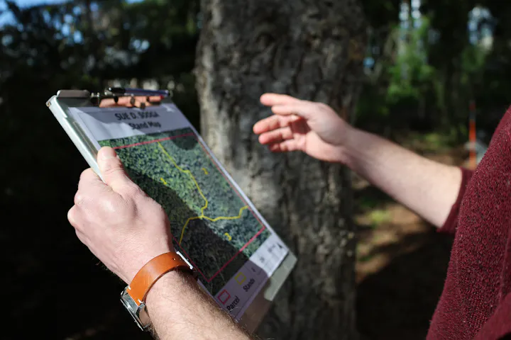 A person holds a clipboard with a detailed map, gesturing while standing near a tree in a forested area.