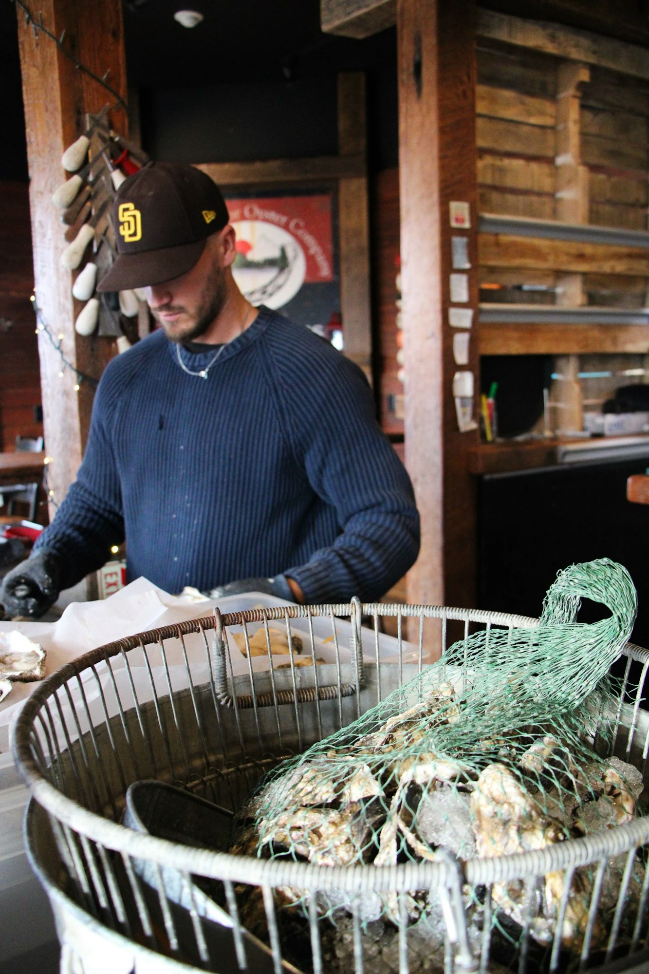 A person in a cap shucking oysters near a basket filled with ice and shellfish.