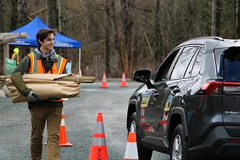Person in a safety vest carries items to a parked car at a roadside setup with cones and a blue tent.