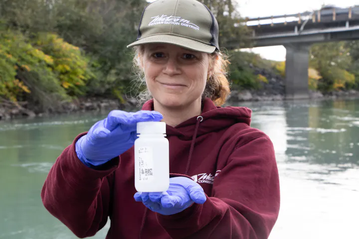 A woman in a sweatshirt and cap holds a sample bottle by a river, wearing gloves and smiling.