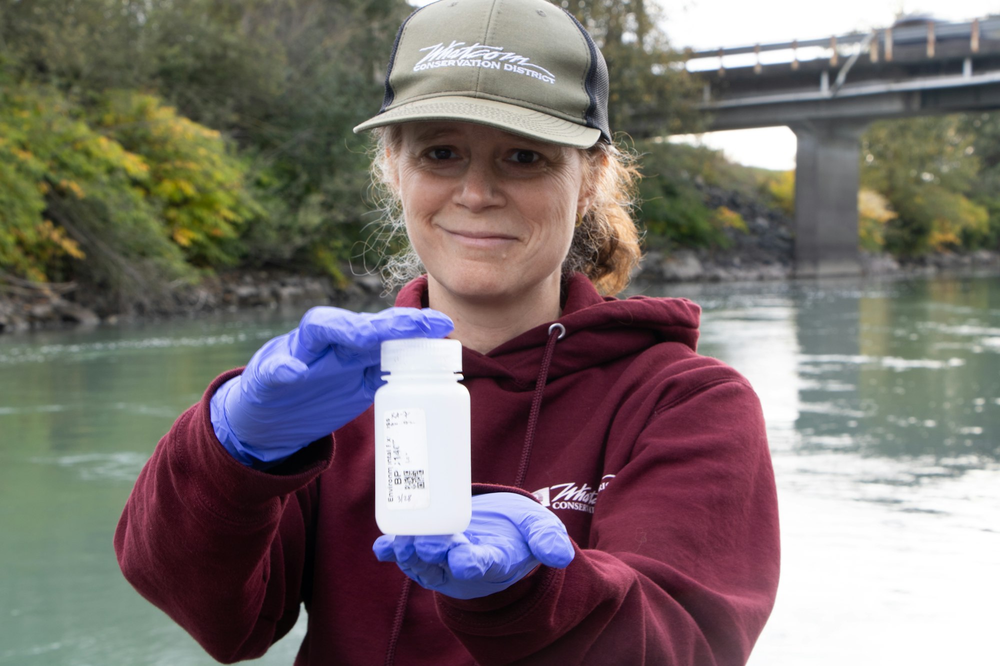 A woman in a sweatshirt and cap holds a sample bottle by a river, wearing gloves and smiling.